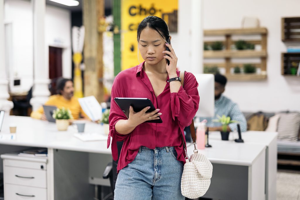  A woman standing in an office, holding a tablet on one hand and answering a phone call on the other.
